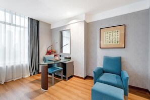 Ein modernes Zimmer mit einem blauen Sessel, einem Schreibtisch mit Stuhl, einem großen Fenster mit Vorhängen und einem gerahmten Kunstwerk an der Wand.