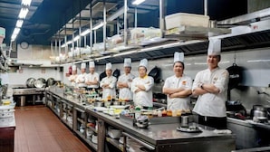A professional kitchen with chefs standing in a row, stainless steel appliances, and various cooking utensils and ingredients on the counters. - Sports International Hotel (Kunshan)