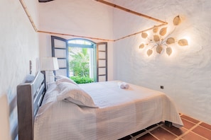 Une chambre à coucher avec un lit, une table de chevet, une lampe et une vue sur la verdure, visible à travers une porte ouverte.