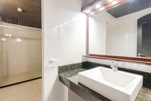 Un baño moderno con cabina de ducha de vidrio, un lavamanos blanco sobre encimera de mármol y un espejo grande con marco de madera.