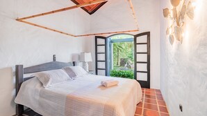 Een slaapkamer met een bed, nachtkastje, lamp en uitzicht op groen via een geopende deur.