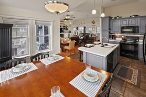 A spacious kitchen with a central island, a dining area with a wooden table, and a view of the backyard through large windows. - Oceanview Home w/ Private Pool, Elevator, Hot Tub, Media Room, Game Rooms (Salvo)