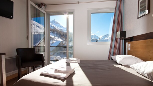 A hotel room with a bed, a chair, a window with a mountain view, and a curtain. - Village vacances Neaclub - La Pulka (Valloire)