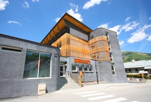 A modern building with a wooden slatted facade, a sign reading "Village of La Follette," and a mountain backdrop. - Village vacances Neaclub - La Pulka (Valloire)