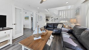 A modern living room with a wooden coffee table, a black leather sofa, a television, and a ceiling fan. - 2-Min Walk to Holmes Beach - Walk to Shops & Dining - Quiet Tropical Townhouse (Holmes Beach)