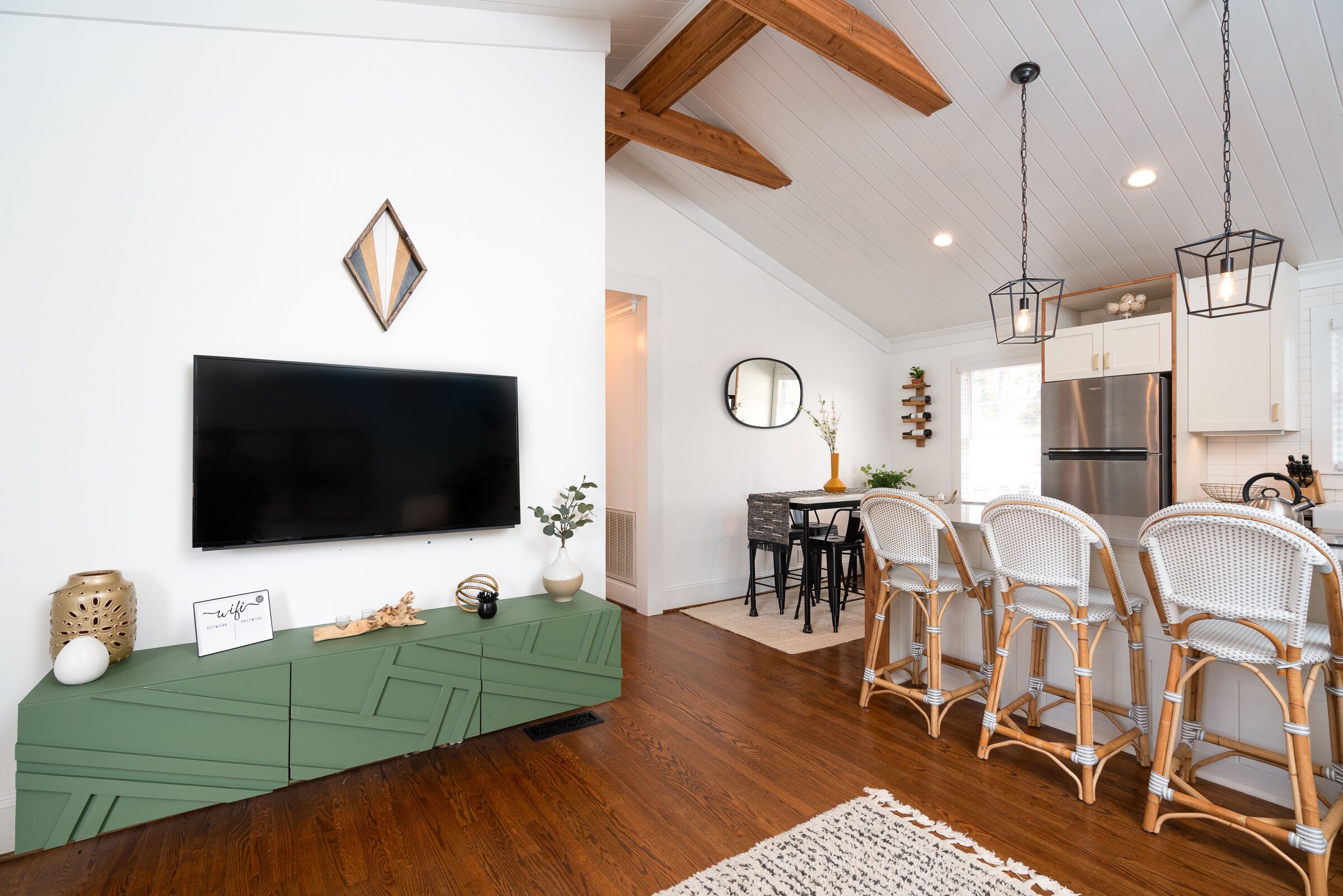 A modern kitchen with a large flat-screen TV, wooden ceiling beams, and a dining area with wicker chairs.