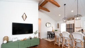 A modern kitchen with a large flat-screen TV, wooden ceiling beams, and a dining area with wicker chairs. - Boho-Chic South CLT Oasis ★ Custom Home ★ Sleeps 6 (Charlotte)