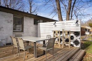 Een terras buiten met een tafel en stoelen, een wit hekwerk met decoratieve details, en een bakstenen gebouw op de achtergrond.