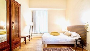 A bedroom with a bed, a wooden wardrobe, a chair, a bedside table, and a window with curtains. - LUXURY UFFIZI - Three Bedroom Apartment, Sleeps 12 (Florence)