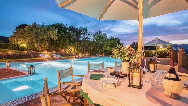 Un área de comedor junto a la piscina, con una mesa lista para comer, rodeada de sillones de descanso y sombrillas.
