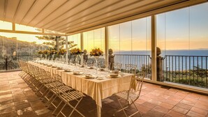 Une longue table à manger, dressée pour un repas, entourée de chaises, avec de grandes fenêtres et une vue sur la mer.