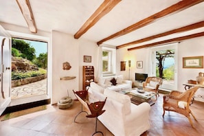 A spacious living room with a stone fireplace, white sofas, and wicker chairs. A large window offers a view of the outdoors. - VILLA MARATEA - Six Bedroom Villa, Sleeps 12 (Maratea)