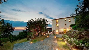 A stone pathway leading to a building with multiple windows, surrounded by lush greenery and a view of the sea. - VILLA MARATEA - Six Bedroom Villa, Sleeps 12 (Maratea)