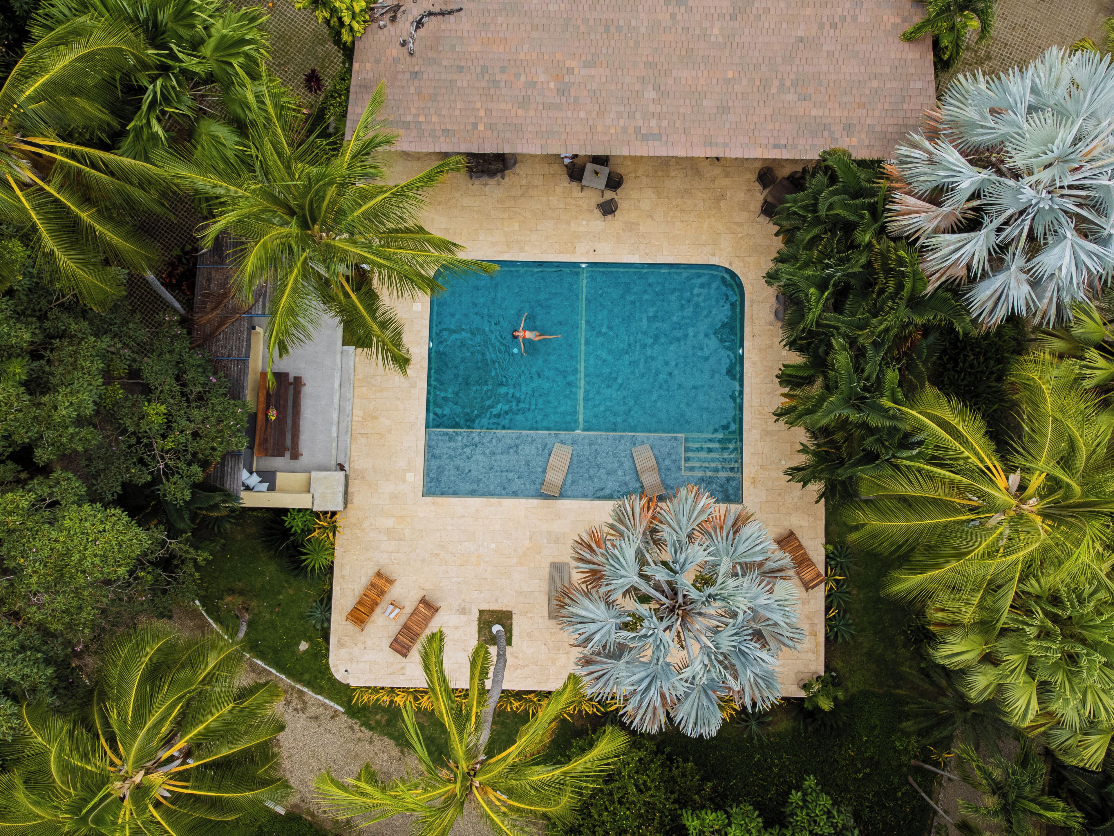 An aerial view of a pool area with a person swimming, surrounded by palm trees and lounge chairs.