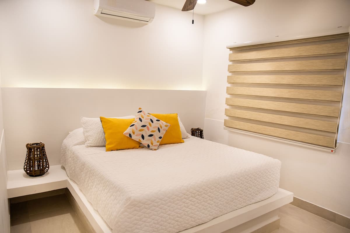 A bedroom with a bed, bedside tables, a wall-mounted air conditioner, and a window with blinds.