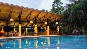 Uma área de piscina com um pergolado de madeira, espreguiçadeiras e uma piscina transparente.