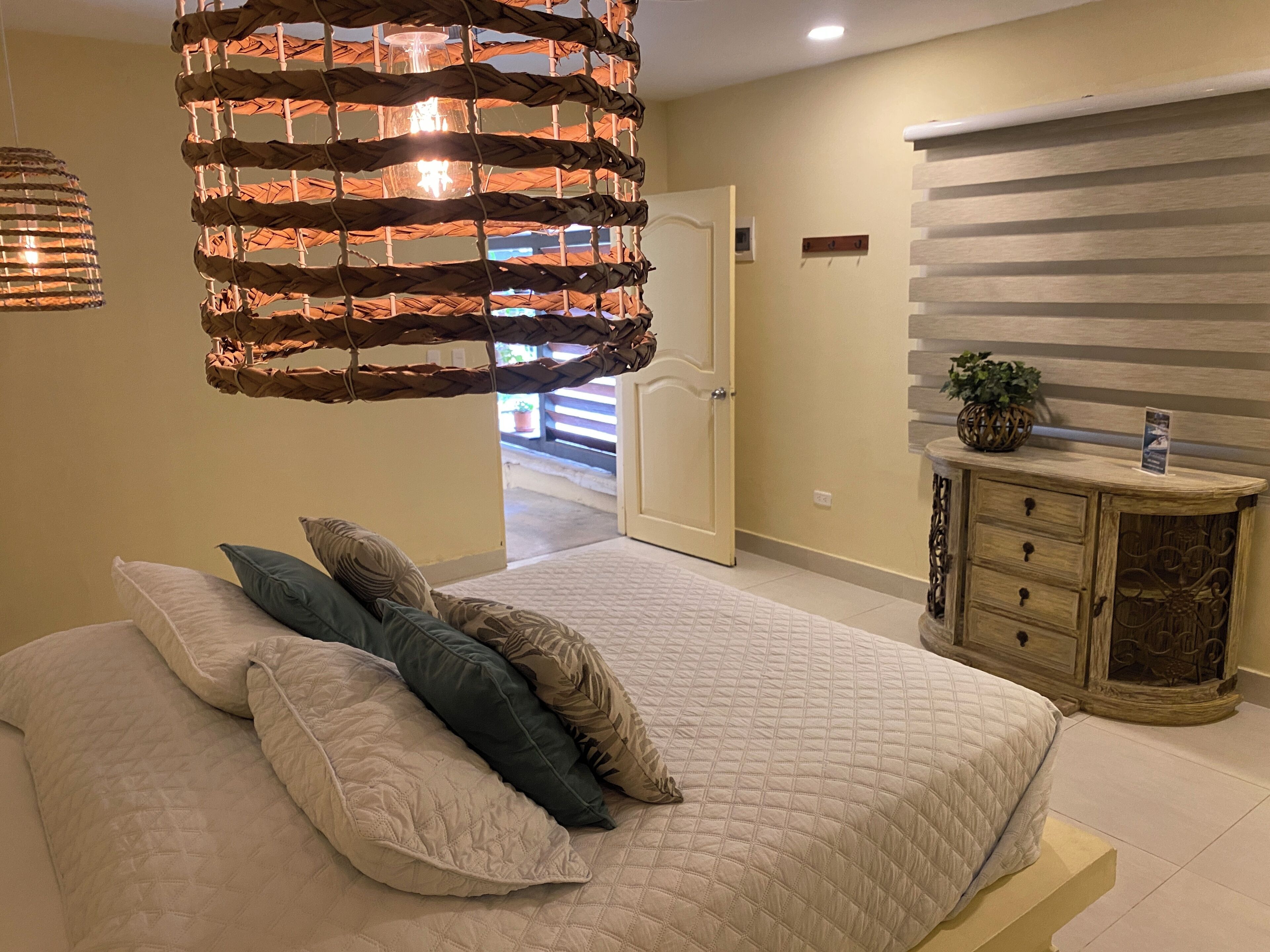 A bedroom with a bed, pillows, a hanging light fixture, a dresser, and a door leading to another room.