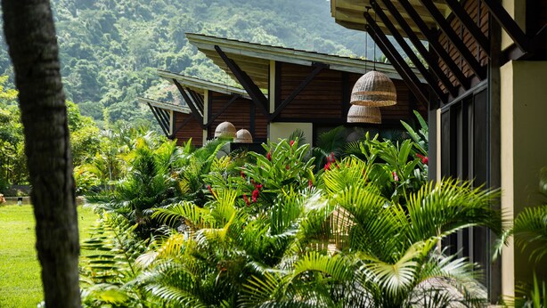 Um edifício com área externa coberta, cercado por vegetação exuberante e montanhas ao fundo.