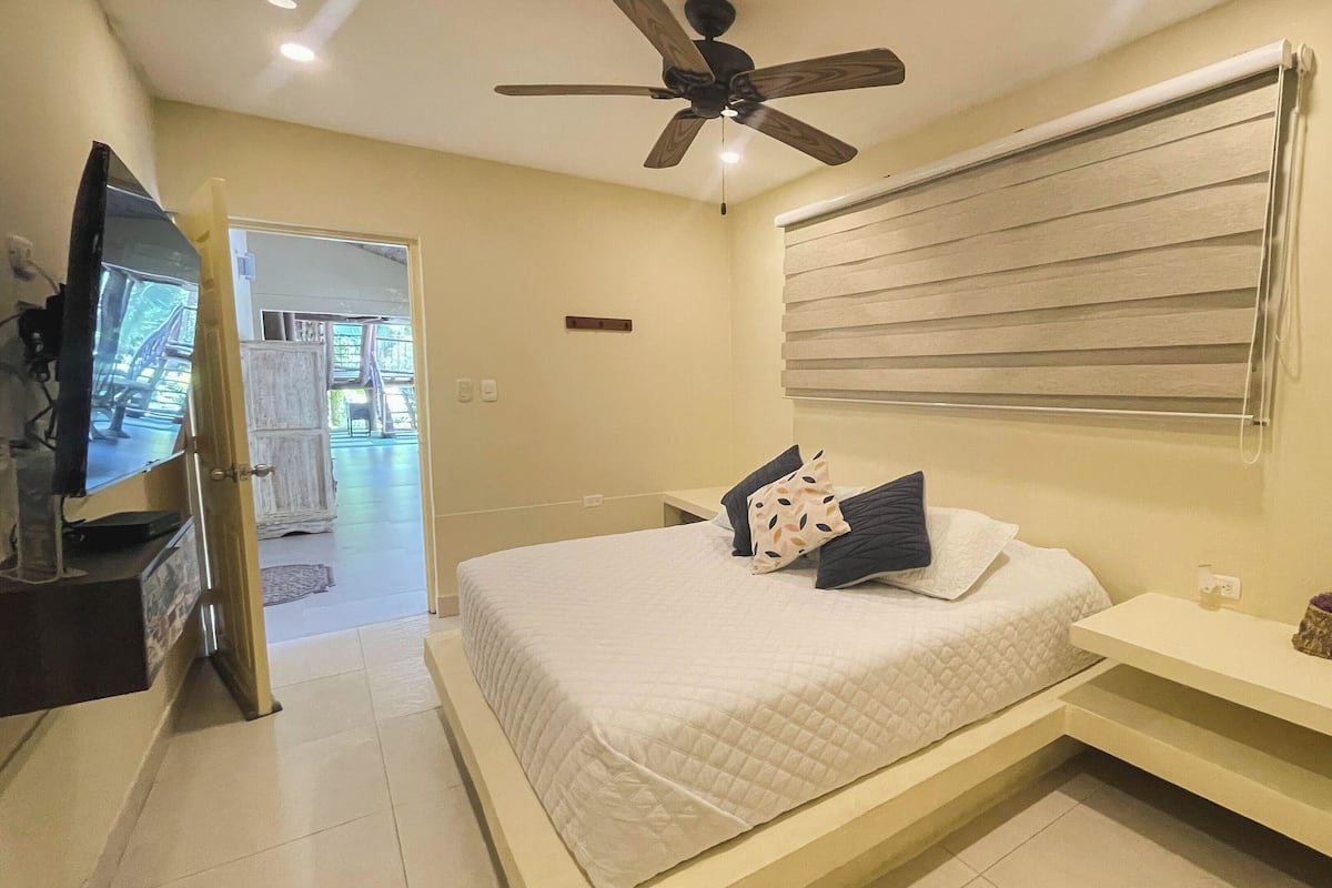A bedroom with a bed, a ceiling fan, a TV, and a window with blinds.