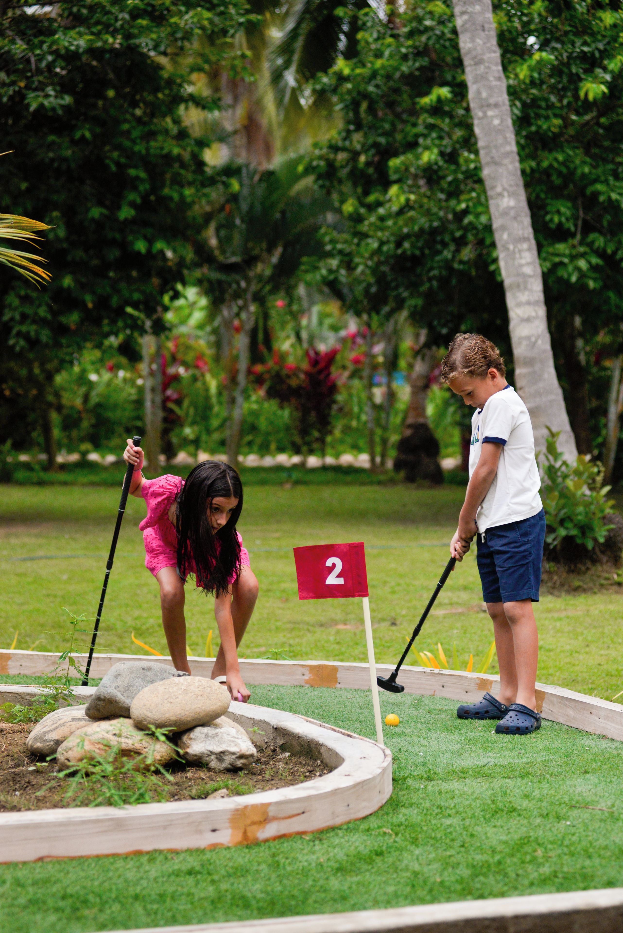 Campo de golf con una bandera roja marcada con el número 2 y una zona de práctica de putts con rocas y césped.
