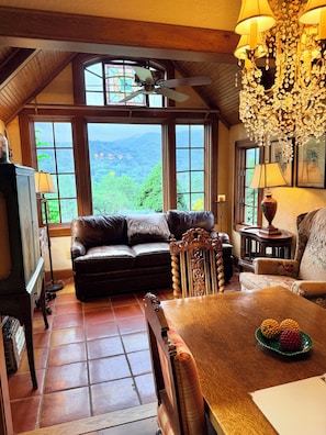 A cozy living room with a leather sofa, a wooden dining table, and a view of a mountainous landscape through large windows. - Elegant Tudor Cottage: Farm Animals, Views, Vintage Decor (Banner Elk)