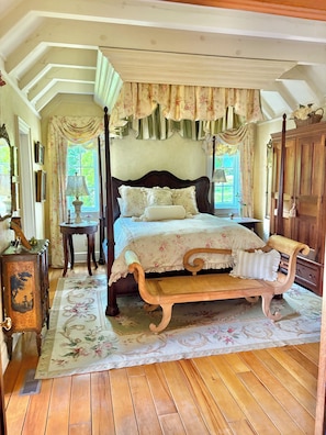 Een hemelbed met bloemenbeddengoed, een houten bankje, een vintage dressoir en een spiegel aan de muur.