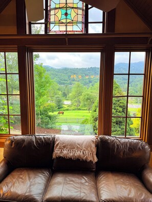 A room with a leather sofa facing a large window with a view of a green landscape and a horse. - Elegant Tudor Cottage: Farm Animals, Views, Vintage Decor (Banner Elk)