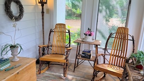 Une pièce chaleureuse avec deux chaises en bois, une petite table avec des livres, une lanterne et une vue sur la verdure à l’extérieur.