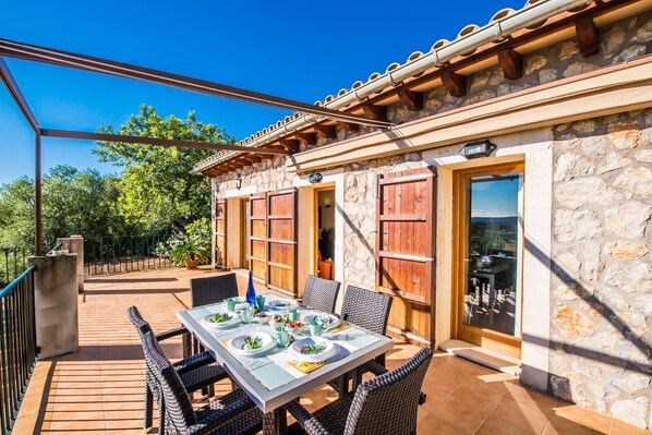 An outdoor dining area with a table set for four, surrounded by wicker chairs, in front of a stone building with wooden shutters and a tiled roof. - Country house in Mallorca Es Puchet with pool (Buger)