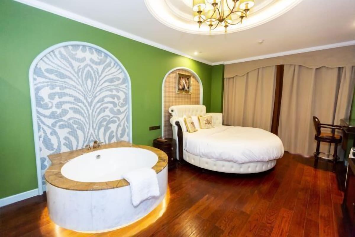 A bedroom with a large round bathtub, a round bed with white bedding, a chandelier, and green walls.