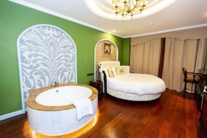 A bedroom with a large round bathtub, a round bed with white bedding, a chandelier, and green walls.