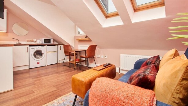 Un espace de vie compact comprenant un coin cuisine, un coin repas et un coin salon avec un canapé et un pouf.