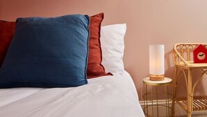 Un lit avec une literie blanche, des oreillers bleus et bruns, une table de chevet avec une lampe et une petite horloge rouge.