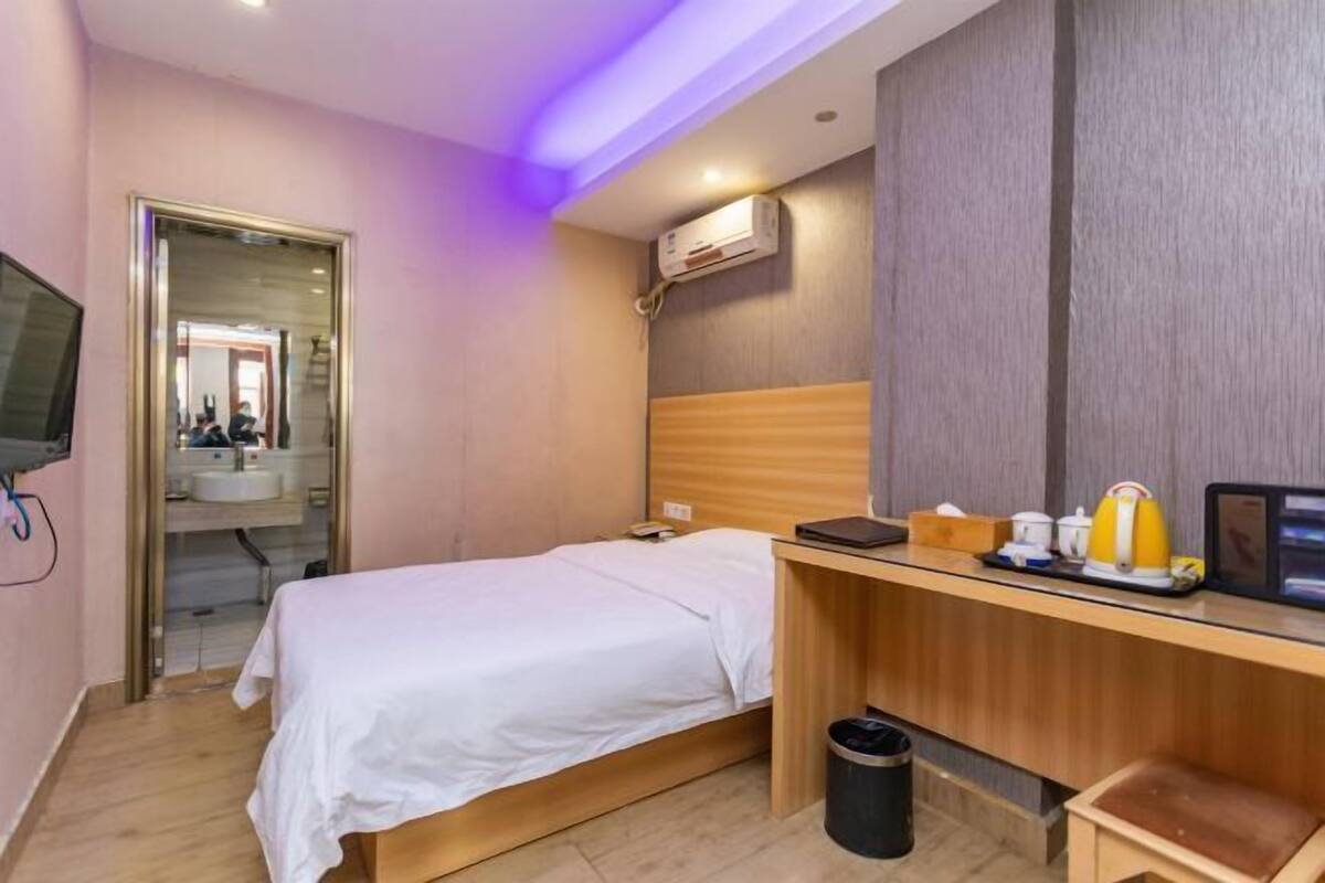 A hotel room with a bed, desk, and a small stool. There is a trash bin and a wall-mounted air conditioner.