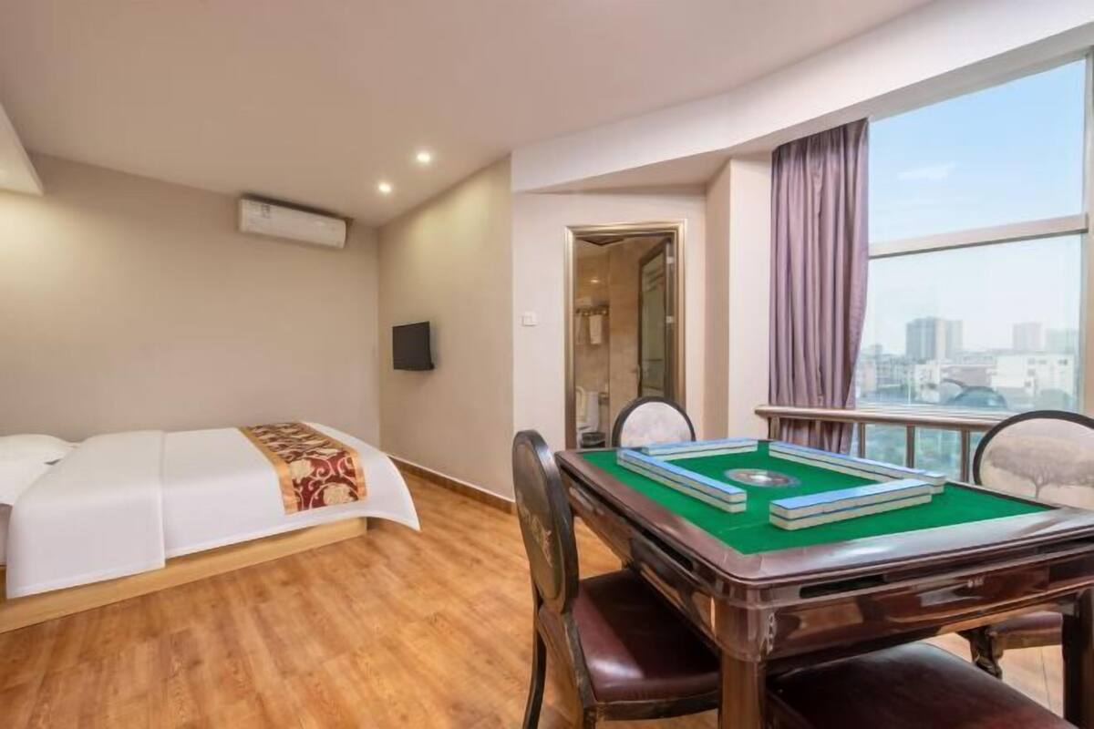 A hotel room with a bed, a green pool table, a chair, and a view of the city through large windows.