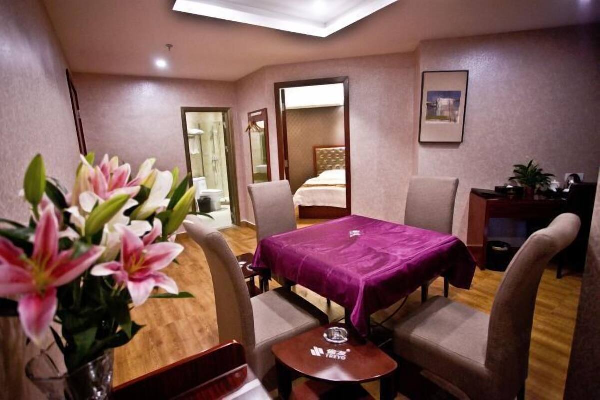 A hotel room with a bed, a table with a purple tablecloth, two chairs, a small table with a glass, and a vase with flowers.