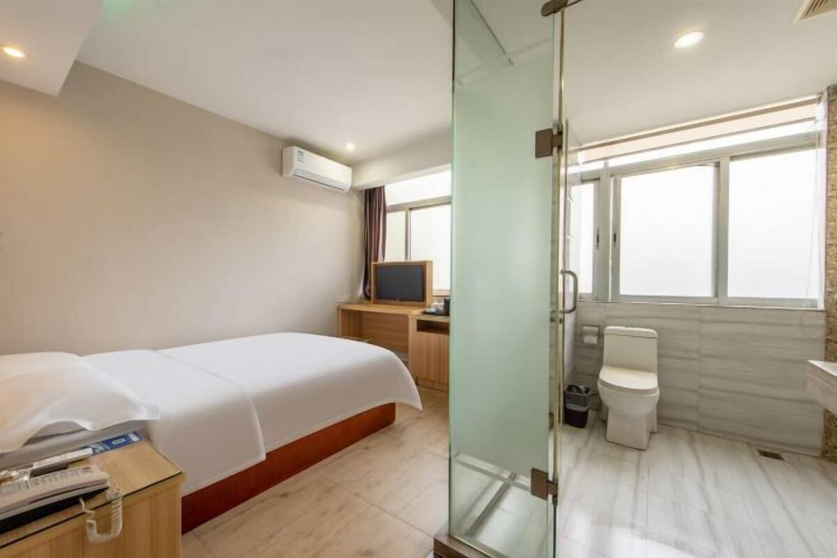 A hotel room with a bed, a desk with a computer, a glass partition, a toilet, and a window.
