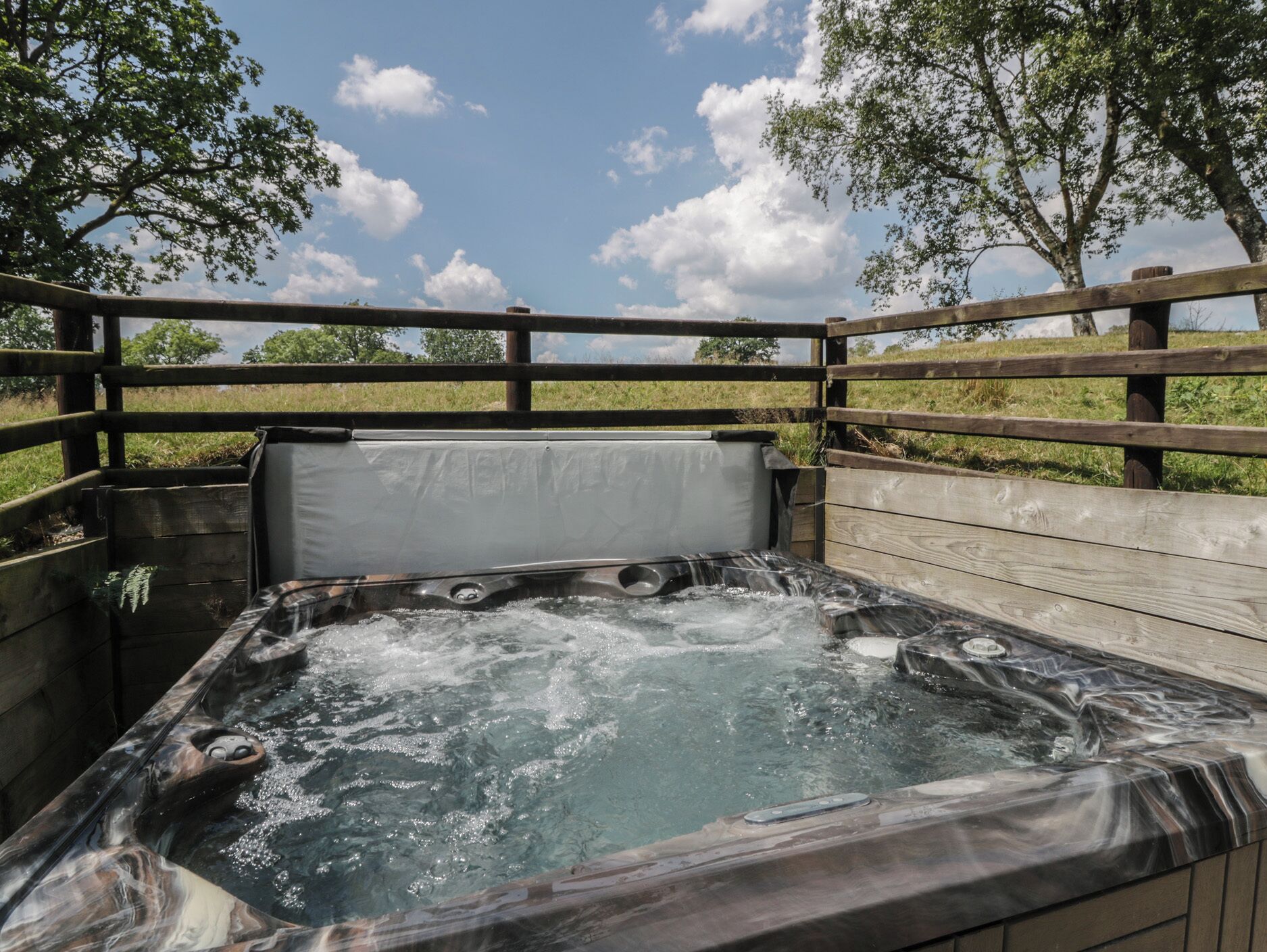 An outdoor hot tub with a wooden deck and metal railings, surrounded by trees and a clear sky.