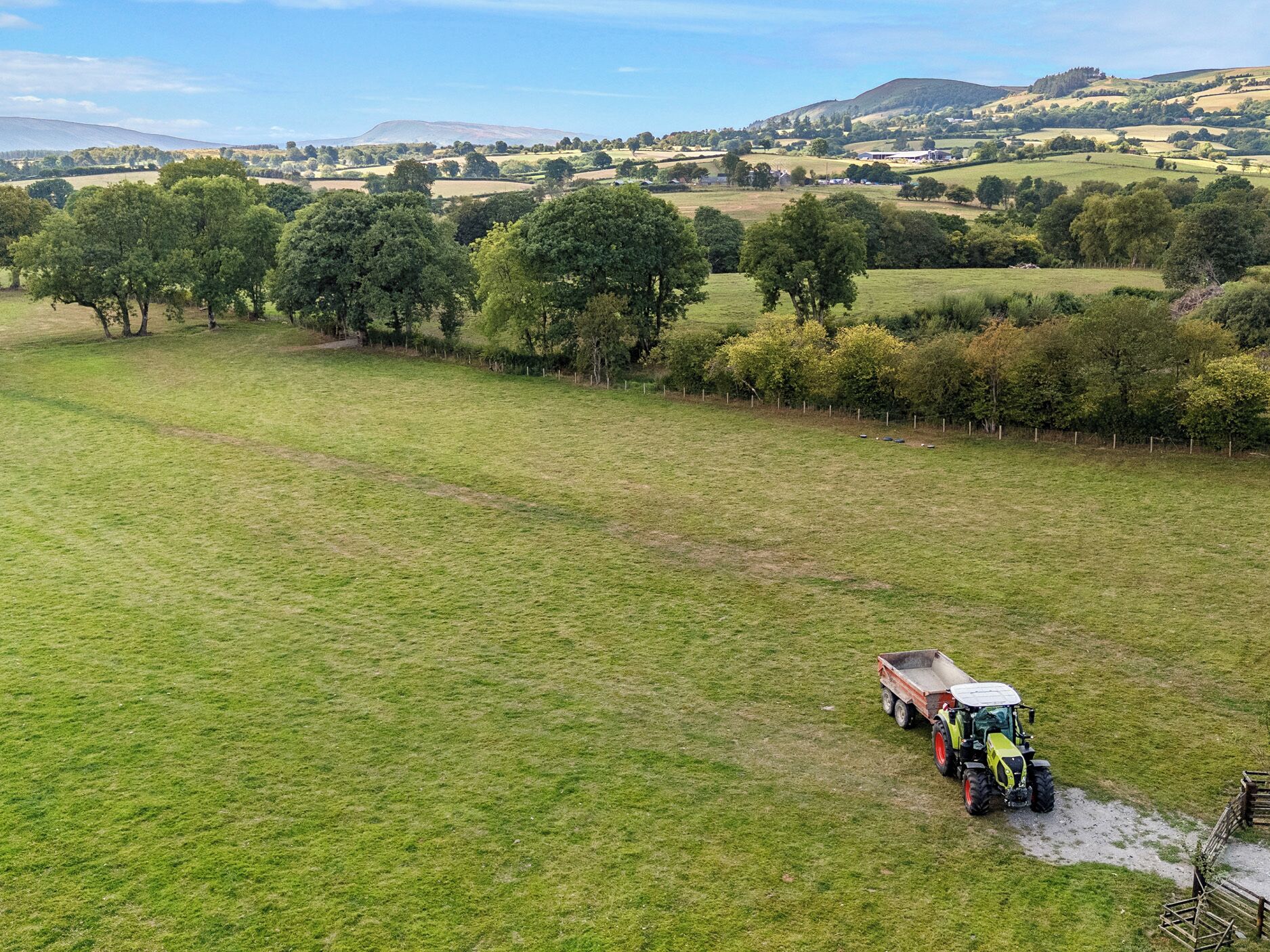 A wide, open field with a tractor and trailer in the foreground, surrounded by trees and rolling hills in the background.