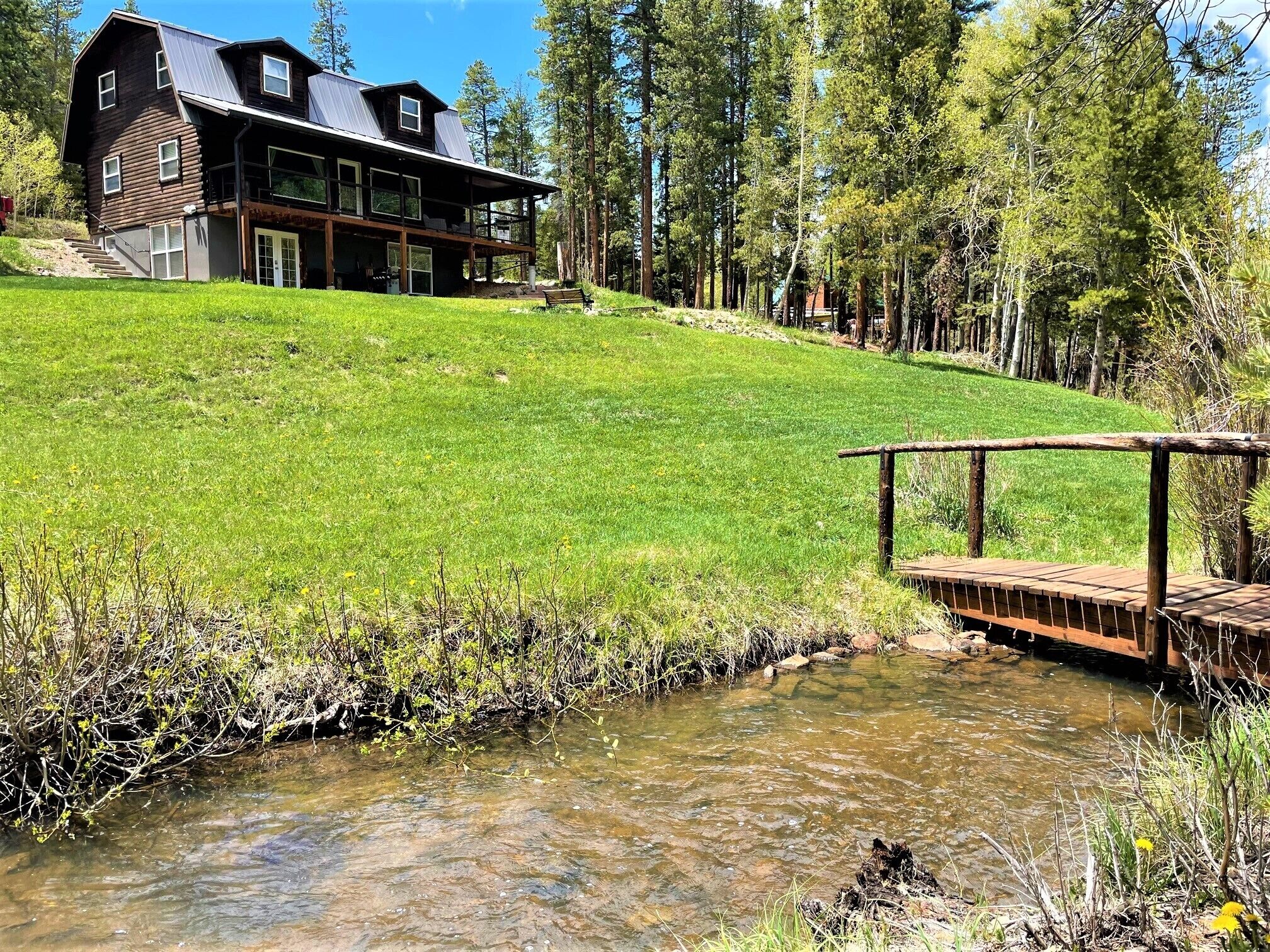 A modern house with a large deck, a small stream, and a forested hillside.