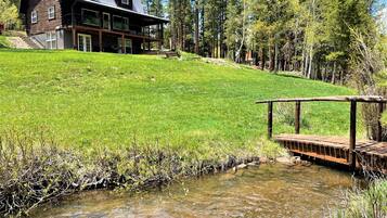 A modern house with a large deck, a small stream, and a forested hillside.