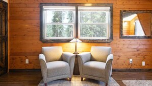 Una habitación acogedora con paredes de madera, dos sillones, una lámpara de mesa y una ventana con vistas a árboles.