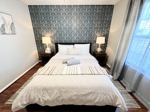 A neatly made bed with a patterned bedspread and pillows, two bedside lamps, a decorative wall with a leaf pattern, and a window with curtains. - Single house: 12guests,4bds/2baths,large backyard. (Jacksonville)