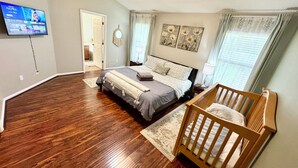 Une chambre avec un grand lit, un berceau en bois, un ventilateur de plafond et un téléviseur fixé au mur.