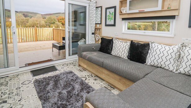 Ein modernes Wohnzimmer mit einem großen Schiebeglasfenster, das auf eine Terrassenfläche führt, einer Couch mit gemusterten Kissen und einer kleinen Holzablage mit Dekorationen.