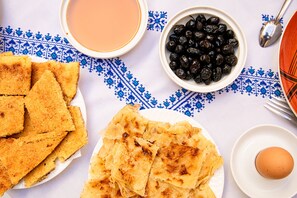 A table with a white cloth featuring blue patterns, a plate of triangular bread pieces, a bowl of black olives, a bowl of pink sauce, and a plate with a single egg. - Riad Sayeda Al Hora (Fes)
