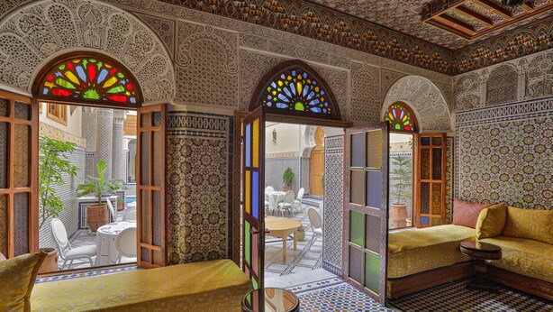 A room with intricate tile work, a sofa, a small table, and a view of an outdoor patio area. - Riad Sayeda Al Hora (Fes)