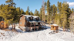 Una cabaña de troncos de dos pisos con un porche y una pequeña cabaña adosada, rodeada de nieve y árboles.