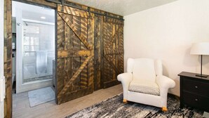 Una habitación con una puerta corredera de estilo granero, un sillón blanco, una mesita de noche negra con una lámpara y una alfombra estampada.