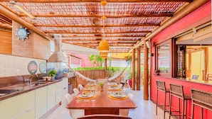 A modern kitchen with a wooden table set for dining, a bar area with stools, and a view of the outdoors. - IT27 Village 3 Suítes Pé na Areia (Lauro de Freitas)